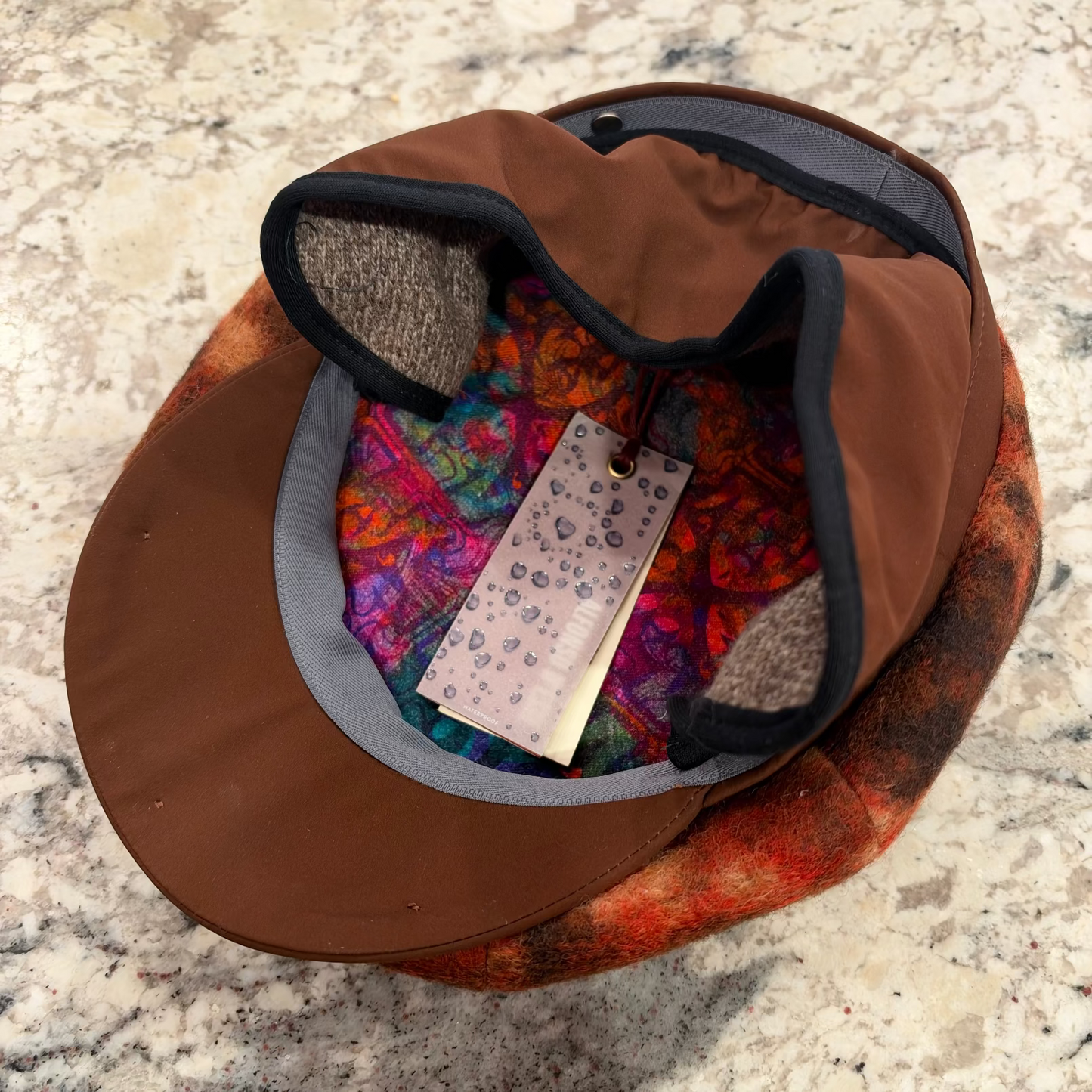 A brown flat-brim cap with a colorful patterned lining and gray trim, photographed from above on a speckled stone surface. A hangtag is visible inside the brim.