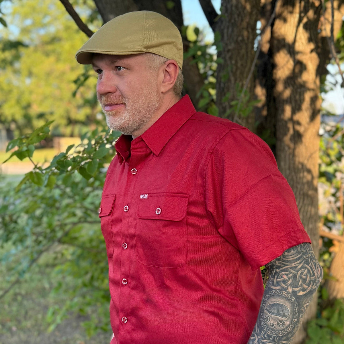 A man wearing a red short-sleeve button-up shirt and a tan flat cap, standing outdoors in front of trees.