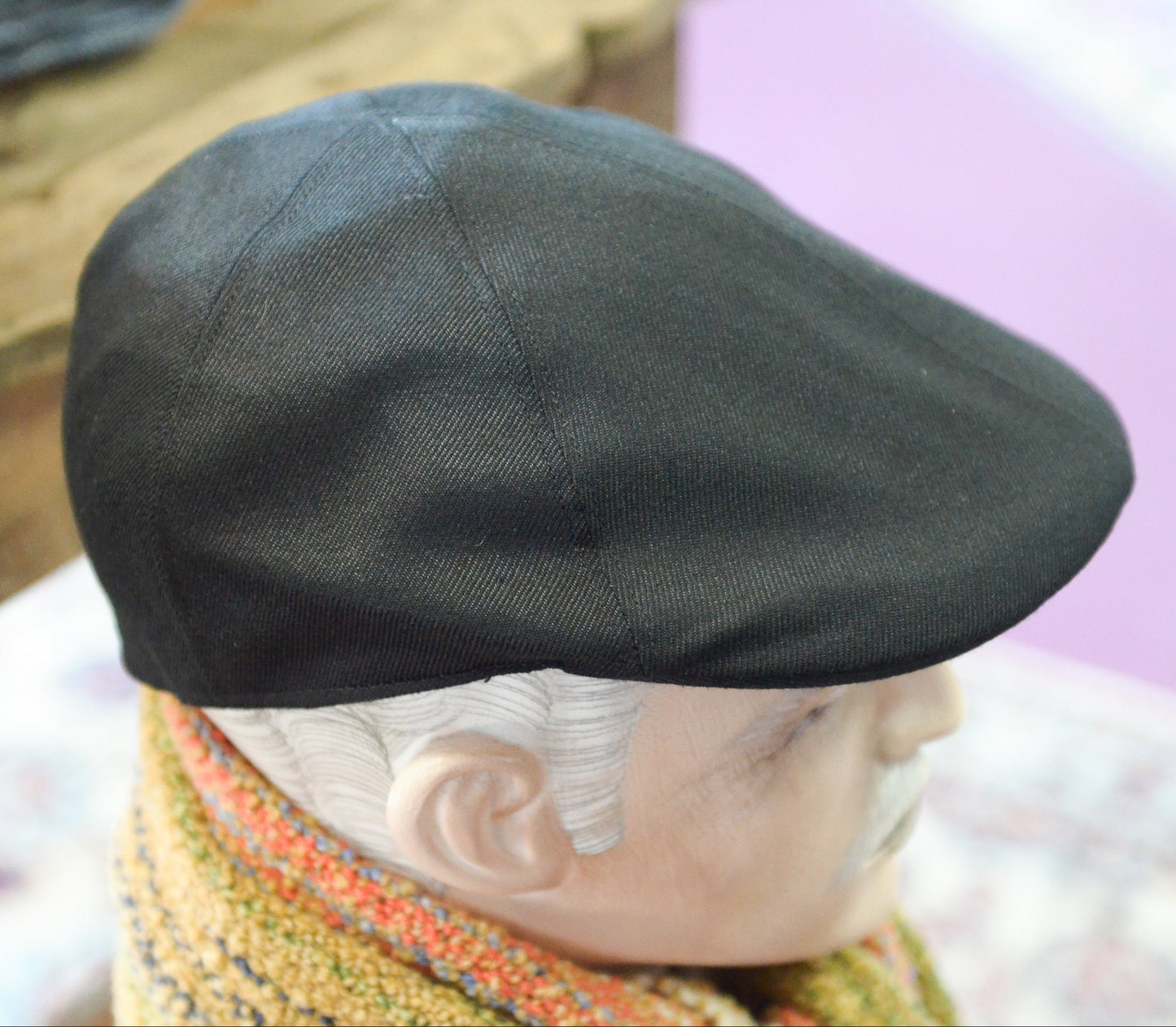 A black flat cap with a textured twill weave, shown on a mannequin head with a colorful knit scarf, set against a softly blurred background.