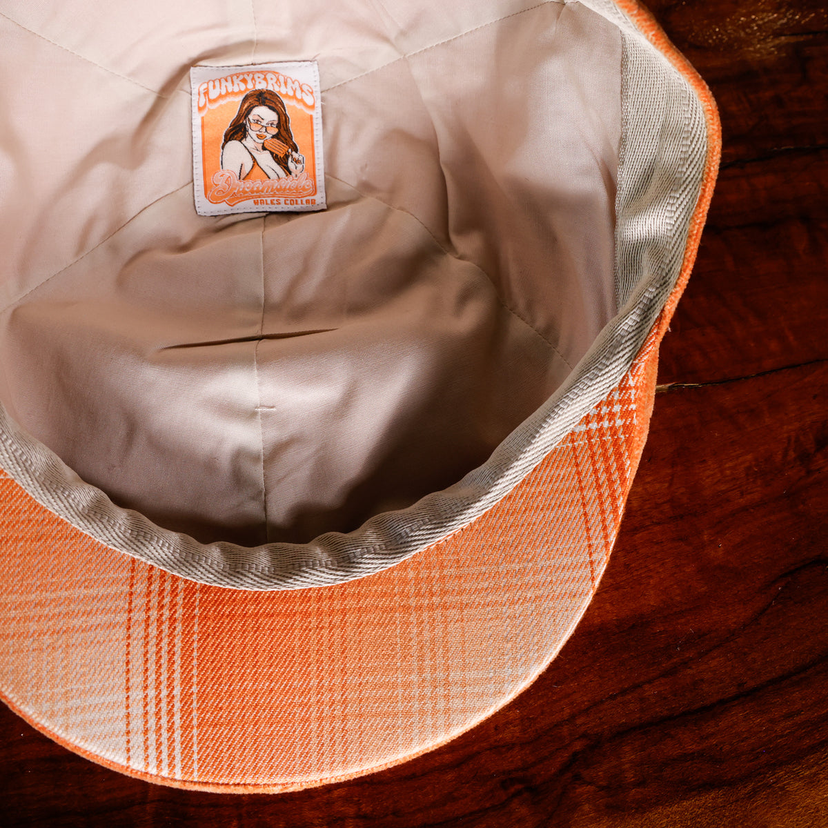 Close-up of the inside of a plaid orange and beige cap resting on a dark wooden surface, showing the interior lining and a sewn-in label.