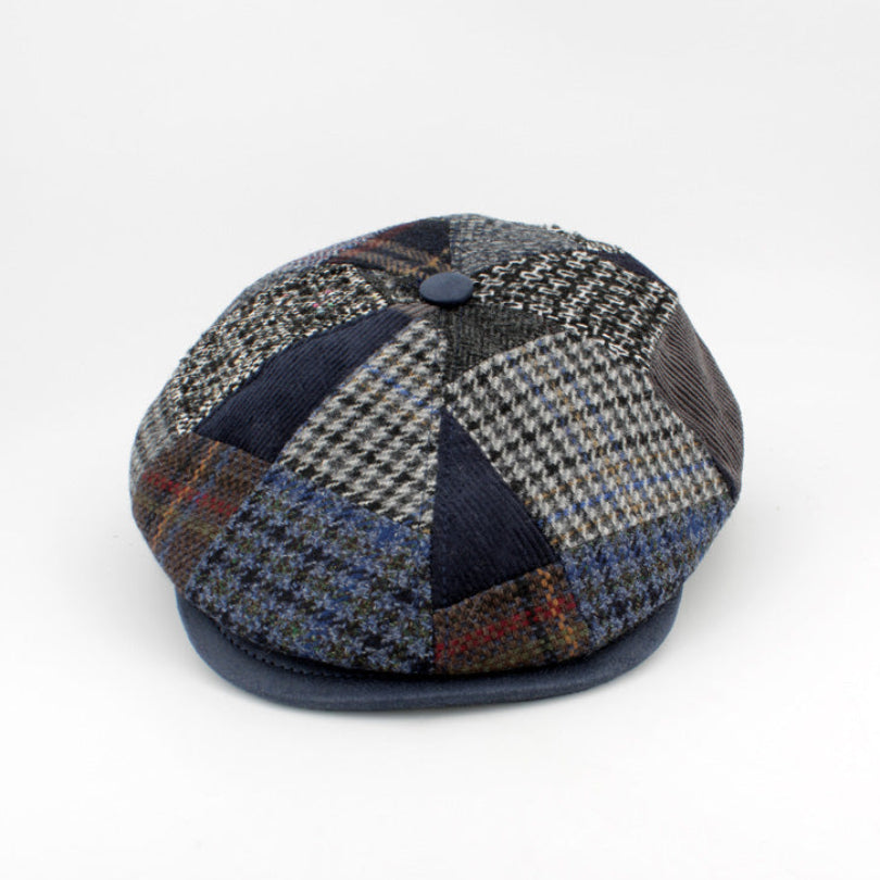 A patchwork flat cap made of various textured tweed and houndstooth fabrics in shades of navy, gray, beige, and brown, with a solid navy blue brim and button.