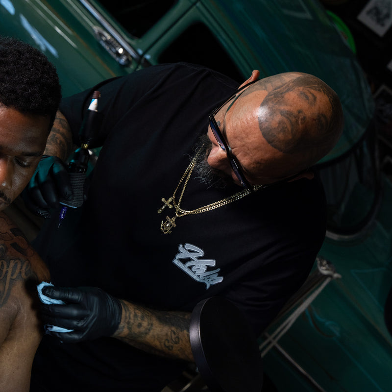 A tattoo artist wearing a black t-shirt with a white script logo, black gloves, and gold necklaces tattoos while working on a client's arm in a dark, teal-toned environment.