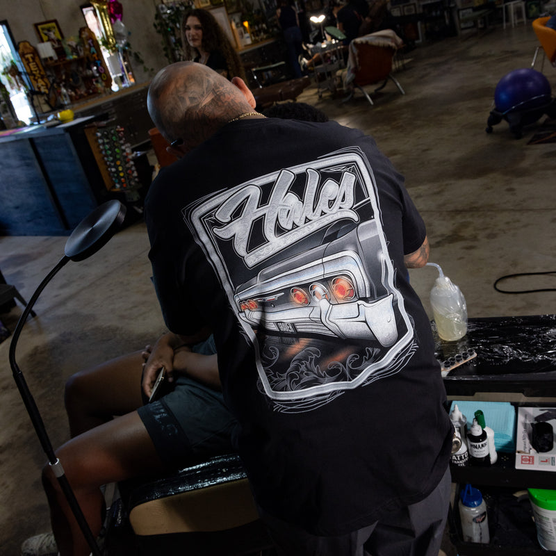 A man wearing a black graphic t-shirt with a large white and gray 'Halo' car design on the back, seen from behind while working at a barber station in a shop.