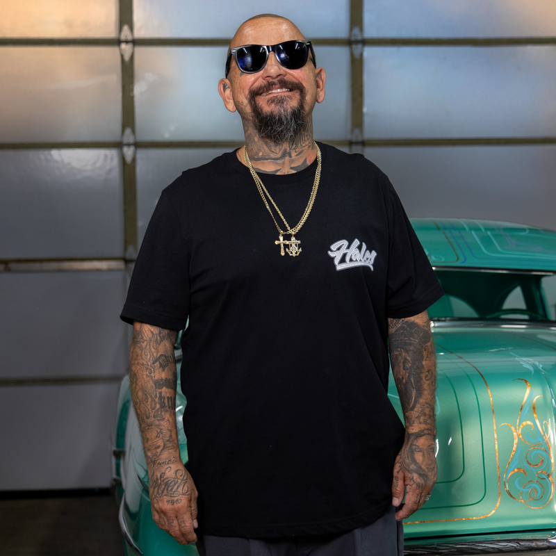 A man with tattoos and sunglasses wearing a black t-shirt with a white 'Hustle' logo, standing in front of a teal classic car.