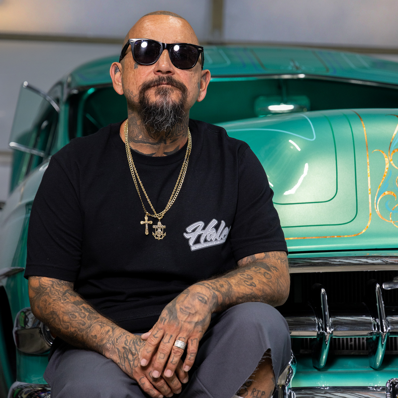 A man with a goatee and sunglasses sits in front of a teal classic car, wearing a black t-shirt with a white script logo and gold chains.