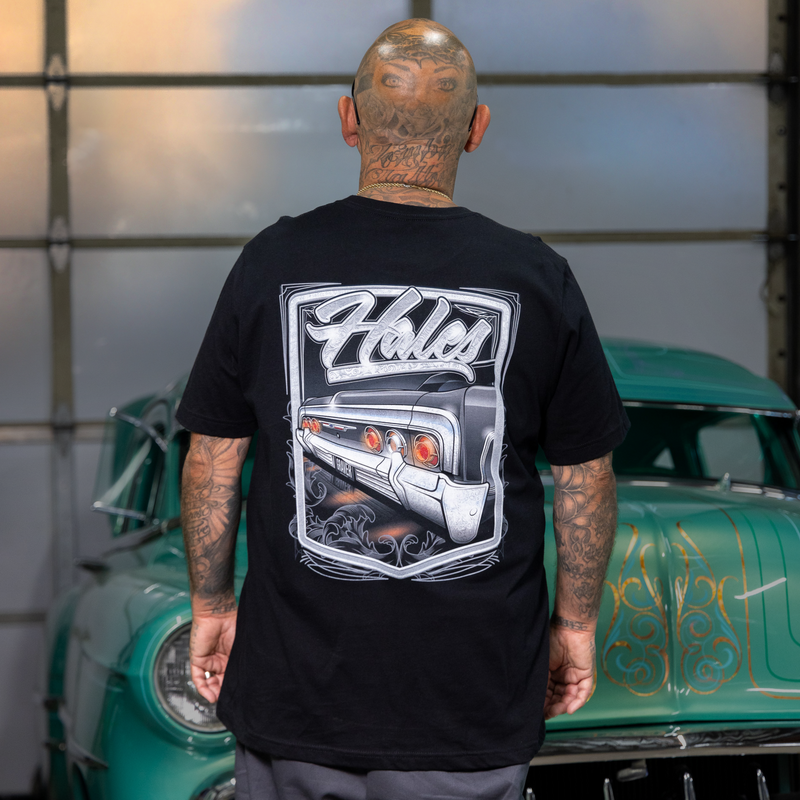 A man with tattoos stands with his back to the camera wearing a black t-shirt featuring a large graphic print of a classic car and the word 'Holes'. A green vintage car is parked in the background inside a garage.