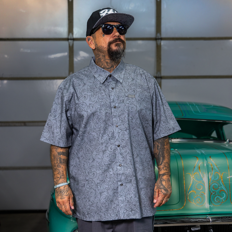 A man with tattoos and a beard wearing a gray short-sleeve button-up shirt with a subtle paisley pattern, a black baseball cap, and sunglasses, standing in front of a vintage green car.