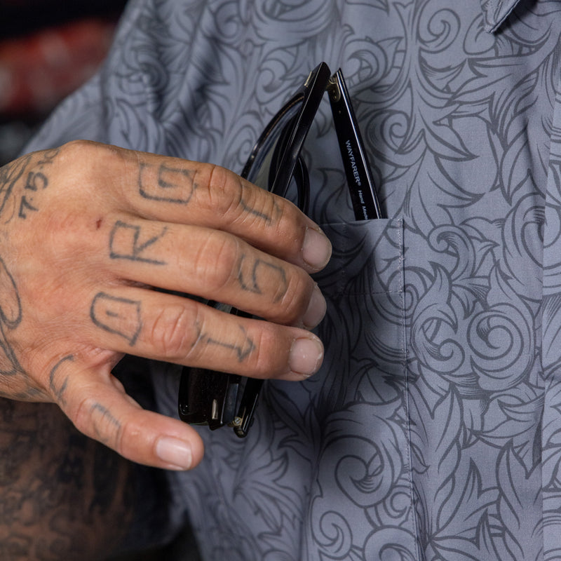 Close-up of a tattooed hand holding a pair of black sunglasses in a gray patterned shirt pocket.