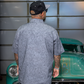 A man viewed from behind wearing a light gray short-sleeve button-up shirt with a dark gray swirling paisley pattern, standing in front of a vintage teal car in a garage.