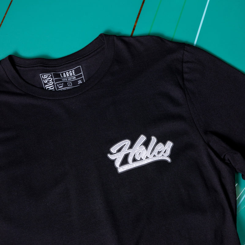A close-up of a black t-shirt featuring a white cursive 'Hales' logo on the chest, laid flat on a vibrant green background.