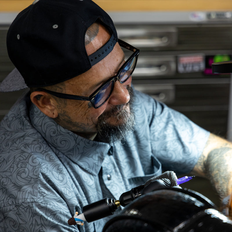 A close-up lifestyle shot of a tattoo artist wearing a black cap, glasses, and a blue-gray patterned shirt, holding a tattoo pen machine in a studio setting.
