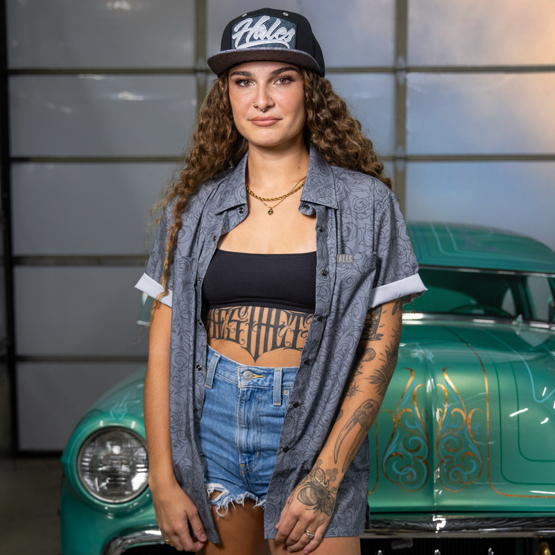 A woman stands in front of a teal vintage car wearing a black and white 'Riesel' baseball cap, a gray patterned button-up shirt over a black crop top, denim shorts, and visible tattoos. The background is a garage interior with metal panels.
