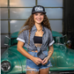 A smiling woman with long curly hair stands in front of a vintage green car, wearing a black and white patterned 'Ulster' baseball cap, a gray patterned button-up shirt tied at the waist over a black crop top, denim shorts, and a gold necklace.