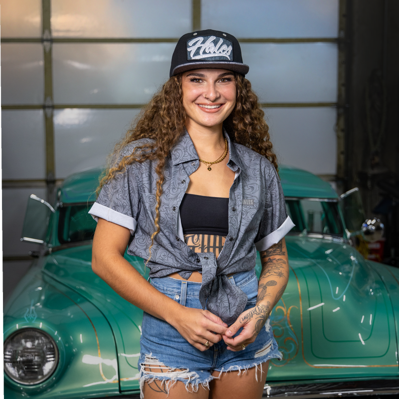 A smiling woman with long curly hair stands in front of a vintage green car, wearing a black and white patterned 'Ulster' baseball cap, a gray patterned button-up shirt tied at the waist over a black crop top, denim shorts, and a gold necklace.
