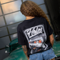 A person viewed from behind wearing a black graphic t-shirt with a large white and gray 'Holes' car design on the back, paired with denim shorts, standing next to a green classic car in front of a glass-paneled wall.