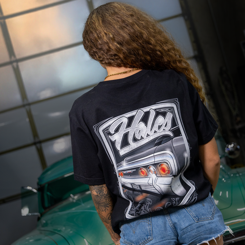 A person viewed from behind wearing a black graphic t-shirt with a large white and gray 'Holes' car design on the back, paired with denim shorts, standing next to a green classic car in front of a glass-paneled wall.