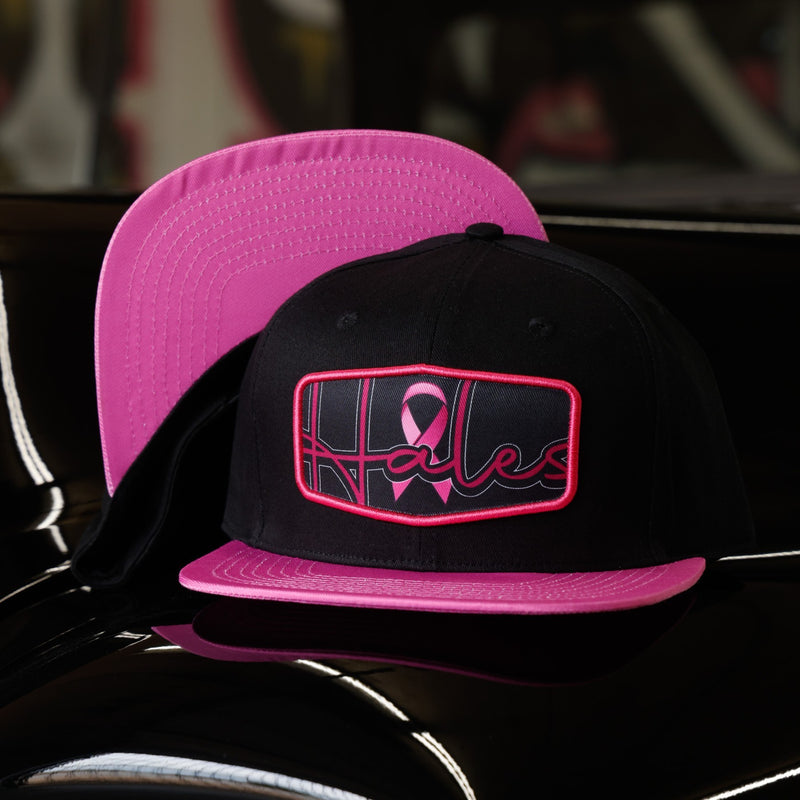 A black and pink snapback cap with a rectangular front patch featuring a pink ribbon and stylized lettering, displayed on a glossy black surface with another matching cap behind it.
