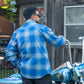 A man wearing a blue and white plaid button-down shirt stands in front of a rustic wooden building next to a blue motorcycle.