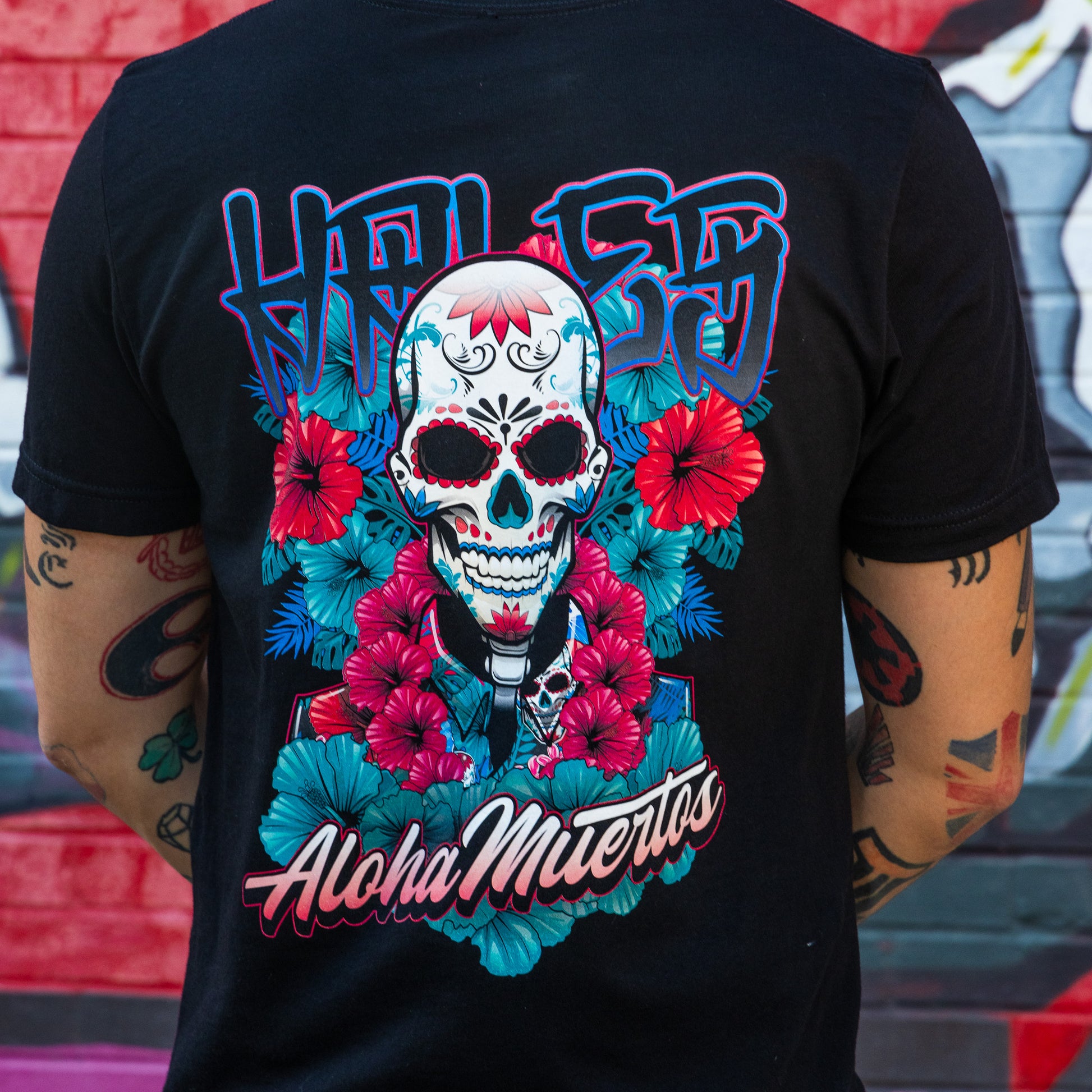 A person wearing a black t-shirt, viewed from the back, featuring a large colorful graphic of a sugar skull surrounded by red flowers and teal leaves, with 'HOLLY' above and 'Aloha Mierros' below.
