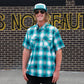 A man wearing a teal and white plaid short-sleeve button-up shirt, sunglasses, and a trucker hat with 'Hikes' on the front, standing in front of a brick wall with large yellow lettering.