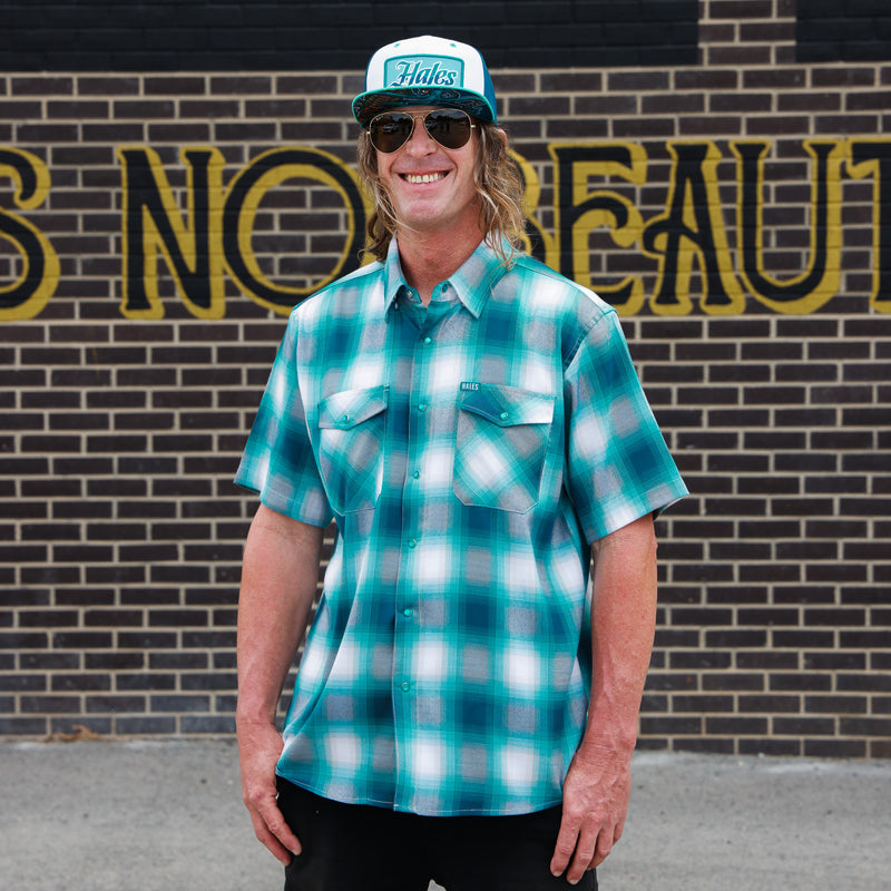 A man wearing a teal and white plaid short-sleeve button-up shirt, sunglasses, and a trucker hat with 'Hikes' on the front, standing in front of a brick wall with large yellow lettering.