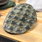 A textured tweed flat cap with a black, beige, and brown houndstooth pattern, resting on a light wooden table.