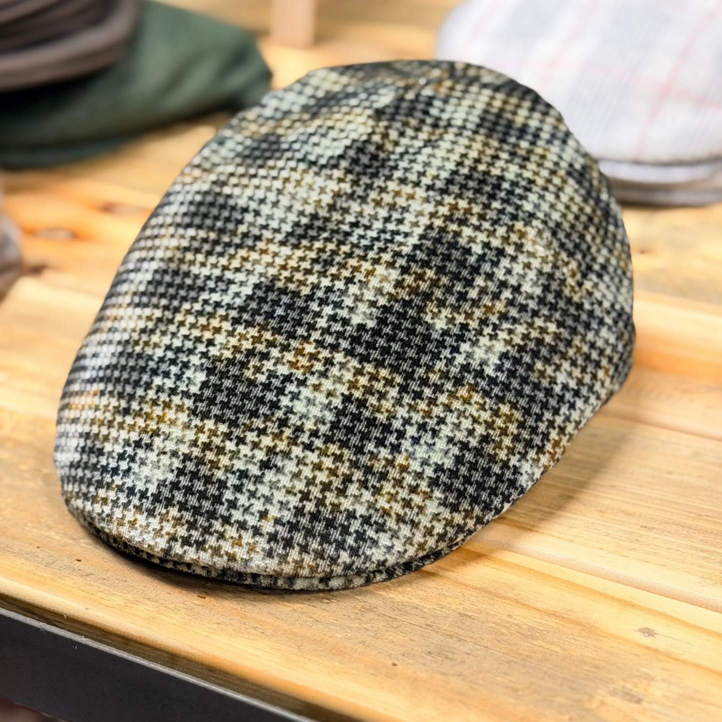A textured tweed flat cap with a black, beige, and brown houndstooth pattern, resting on a light wooden table.