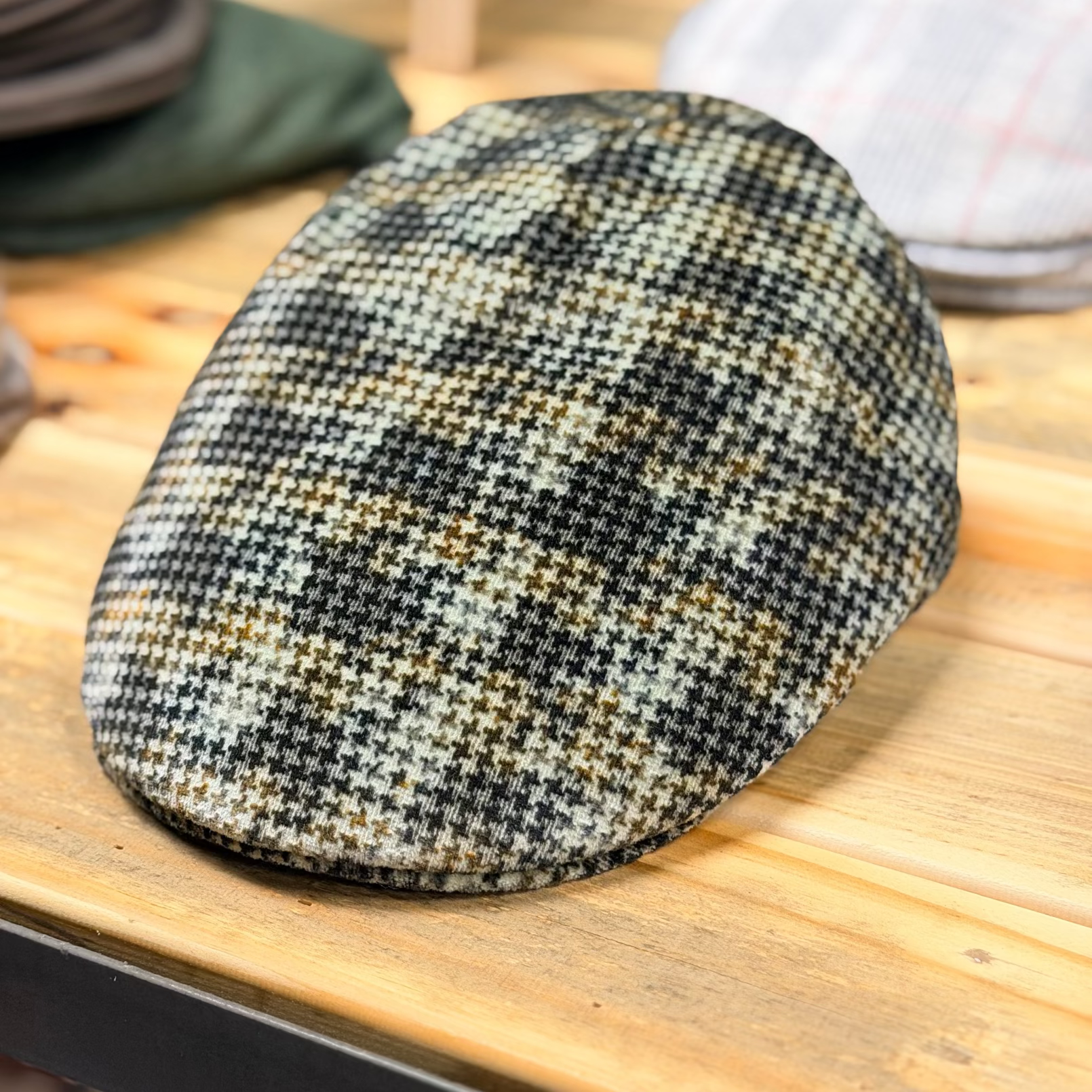 A textured tweed flat cap with a black, beige, and brown houndstooth pattern, resting on a light wooden table.