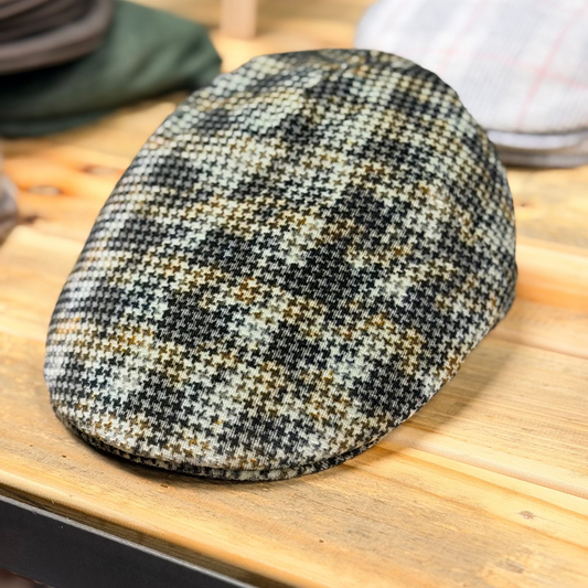 A textured tweed flat cap with a black, beige, and brown houndstooth pattern, resting on a light wooden table.