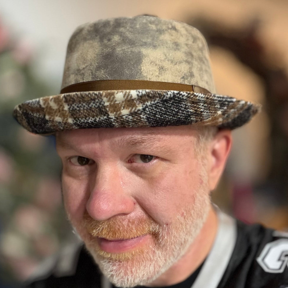 A close-up portrait of a man wearing a textured, distressed camouflage-style fedora hat with a brown band and a patterned brim, set against a blurred indoor background.