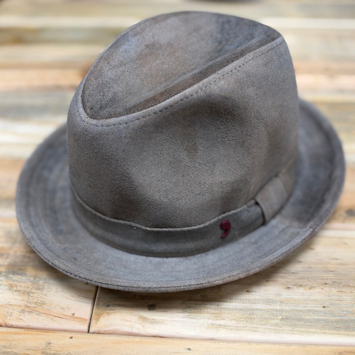 A distressed gray felt fedora hat with a matching band and a small red accent, resting on a rustic wooden plank surface.