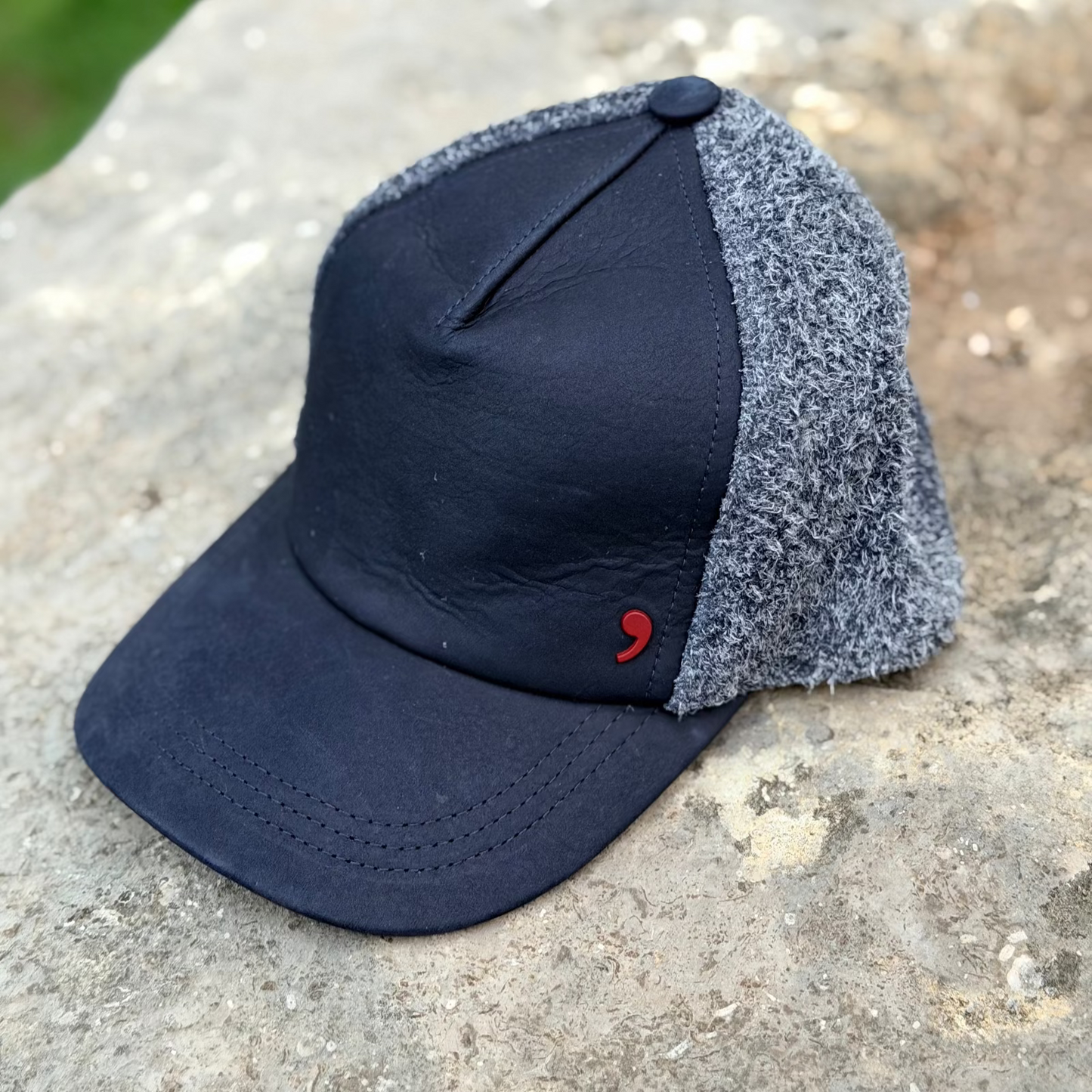 A baseball cap with a dark navy leather front and bill, and a textured gray fuzzy mesh back, resting on a stone surface.