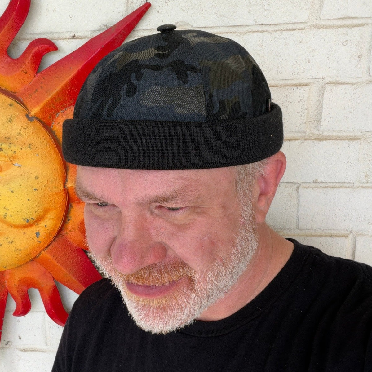 A man with a beard wearing a black camouflage trucker hat with a black knit brim, standing against a white brick wall next to a red and yellow sun wall decor piece.