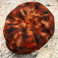 A close-up of a round felt flat cap viewed from above, featuring a mottled pattern of red, orange, brown, and black, with a small red button at the center. The cap sits on a speckled gray and white stone surface.