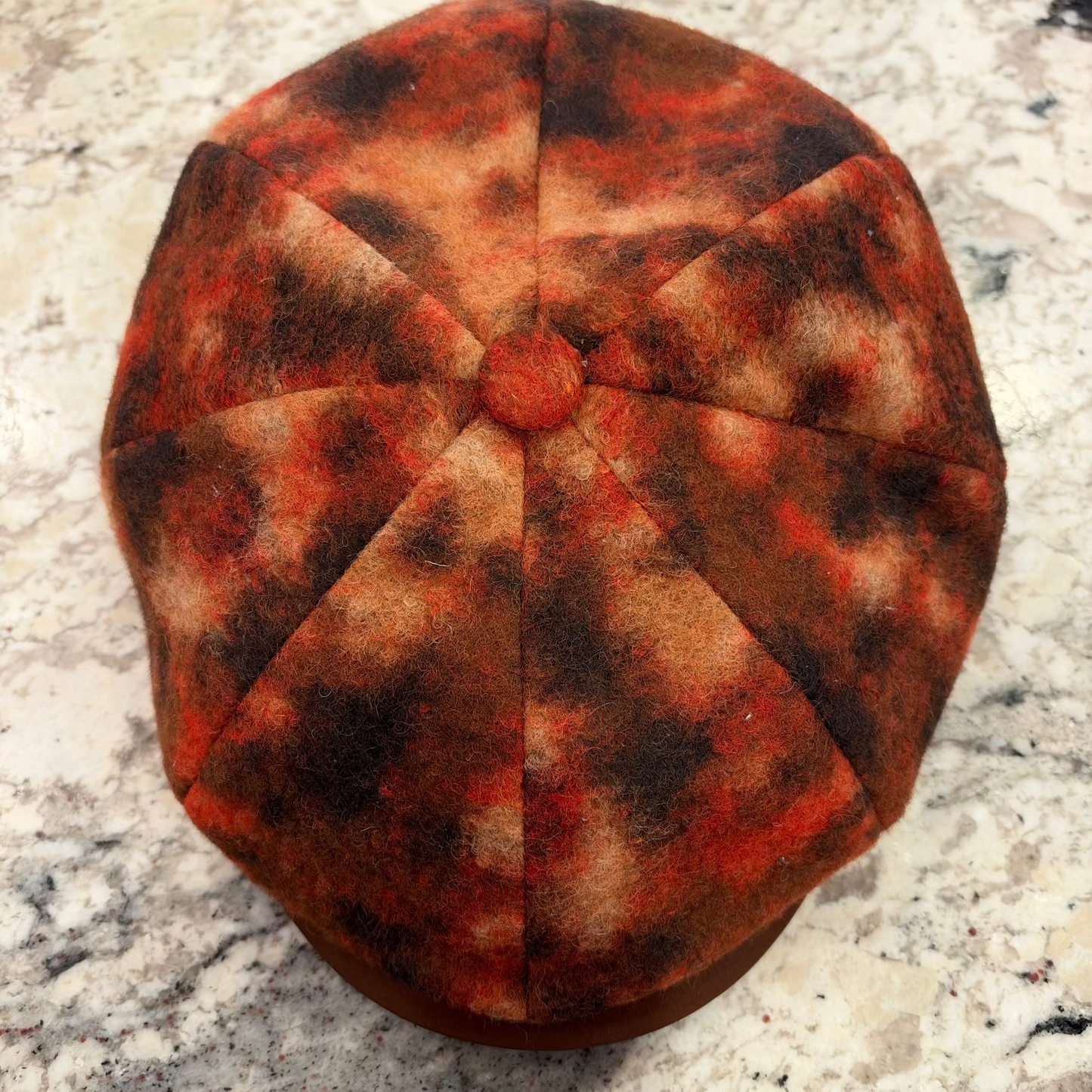 A close-up top-down view of a fabric-covered octagonal cap or hat, featuring a mottled pattern of rust red, orange, and dark brown on a light beige background, set against a speckled stone surface.