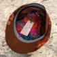A brown flat-brim cap with a colorful patterned lining and a patterned outer fabric, shown from above on a speckled stone surface. A hangtag is attached inside the brim.