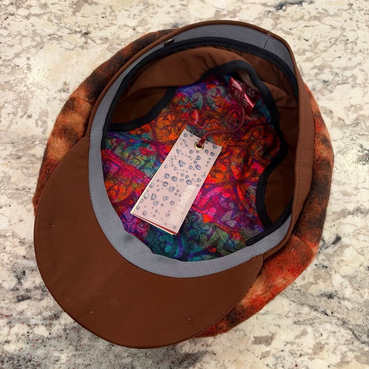 A brown flat-brim cap with a colorful patterned lining and a patterned outer fabric, shown from above on a speckled stone surface. A hangtag is attached inside the brim.