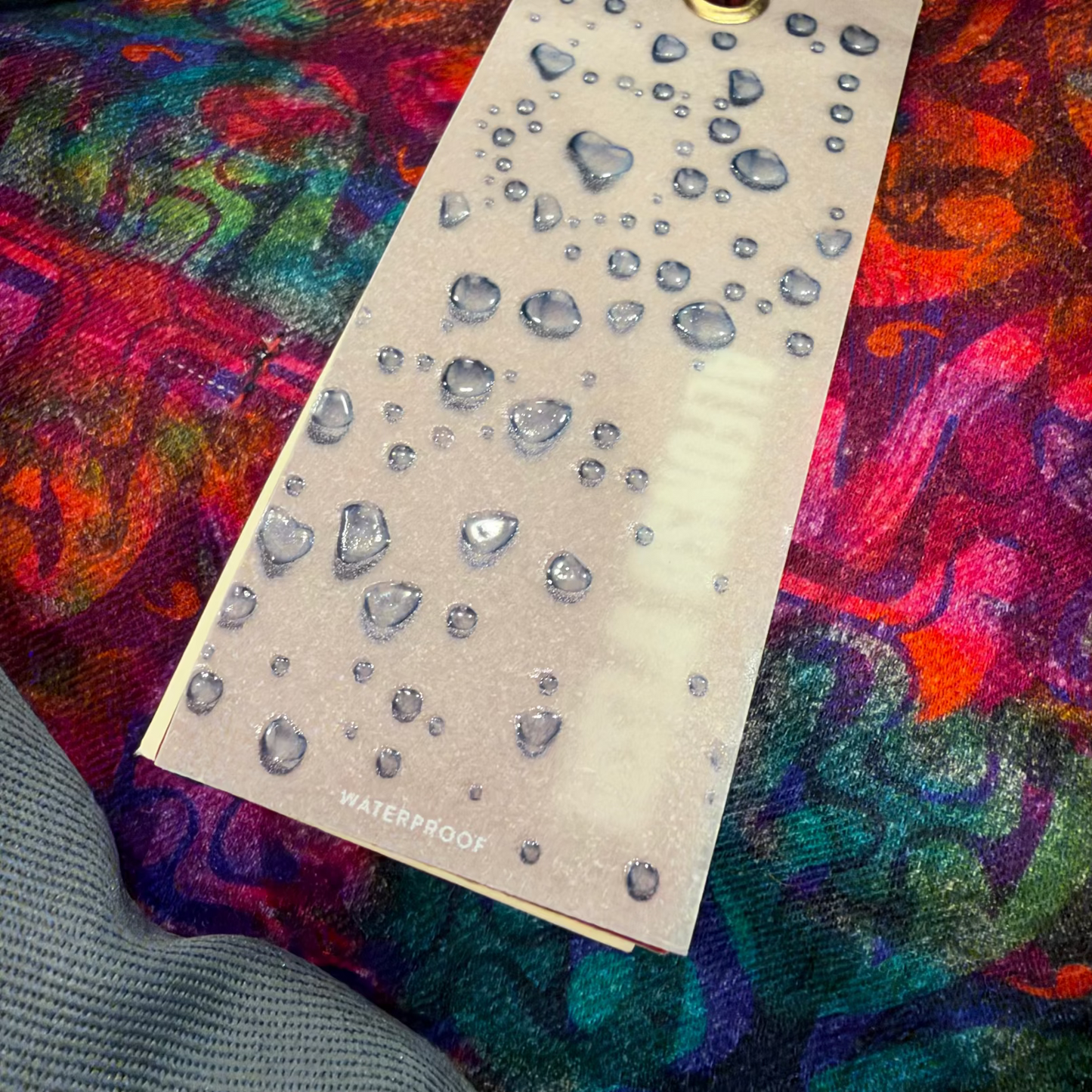 A rectangular beige waterproof test strip with water droplets on it, placed on a highly colorful, patterned fabric background. The word 'WATERPROOF' is printed at the bottom of the strip.
