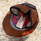 A brown flat-brim cap with a colorful patterned lining and gray trim, photographed from above on a speckled stone surface. A hangtag is visible inside the brim.
