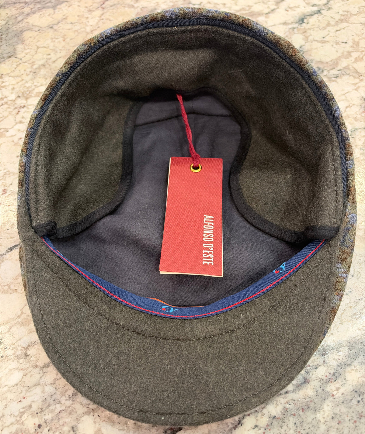 An overhead view of a wool cap's interior, showing a dark olive green wool body, a gray inner lining, and a red hanging tag.