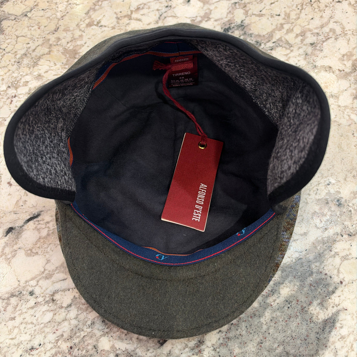 An olive green wool cap with a black felt underbrim and a red interior tag, photographed from above on a speckled granite countertop.