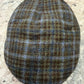 A classic flat cap in a brown, beige, and blue plaid tweed pattern, photographed from directly above on a speckled stone countertop.