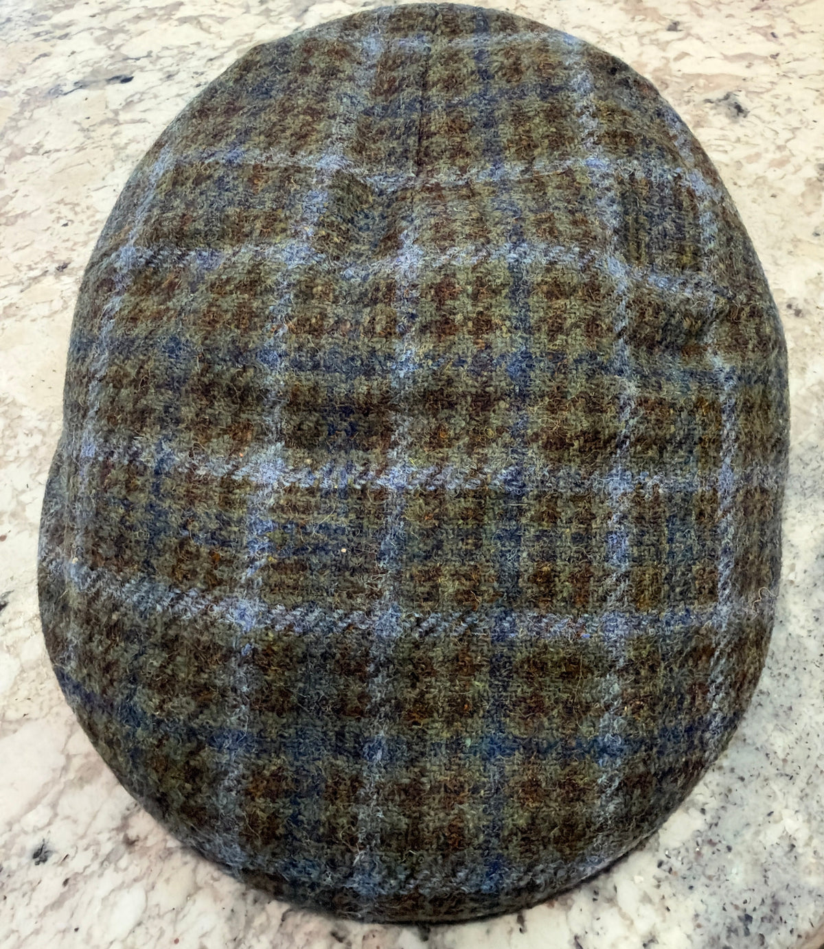 A classic flat cap in a brown, beige, and blue plaid tweed pattern, photographed from directly above on a speckled stone countertop.