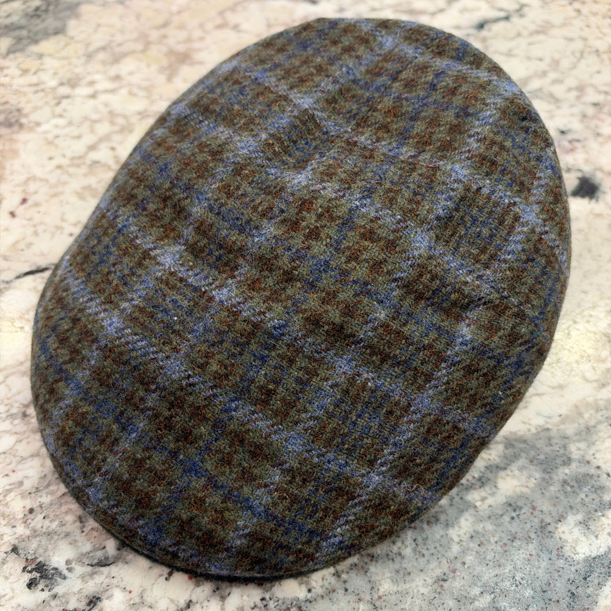 A classic flat cap made of brown and blue tweed with a subtle plaid pattern, resting on a speckled granite countertop.