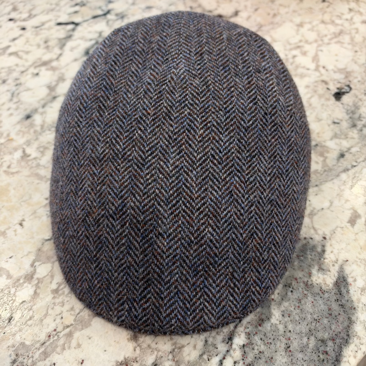 A classic flat cap in a dark brown and gray herringbone tweed pattern, resting on a light-colored stone surface.