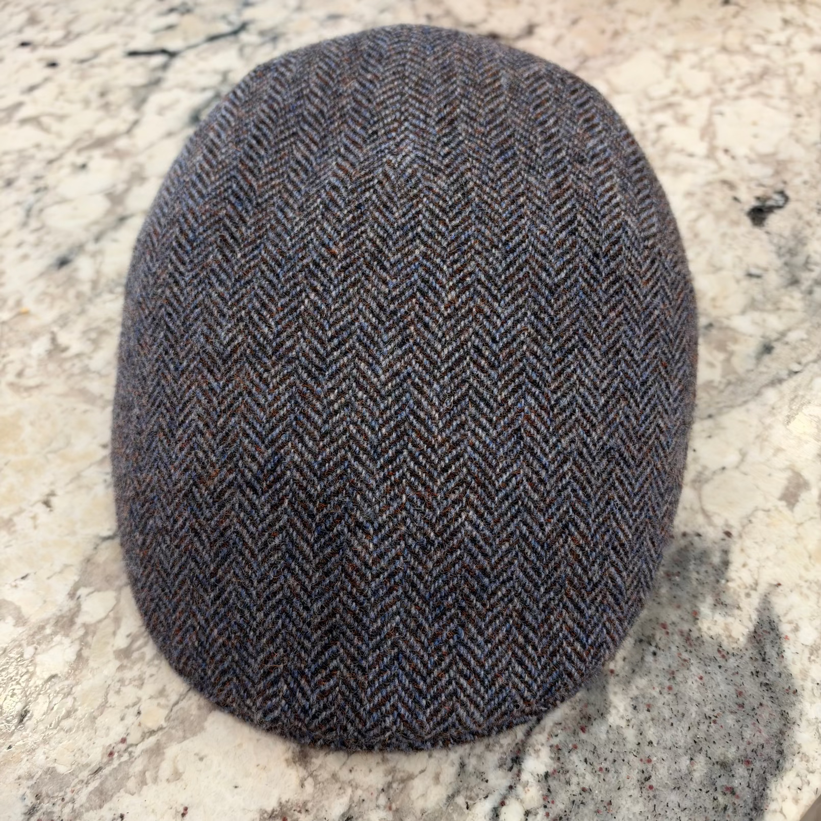 A classic flat cap in a dark brown and gray herringbone tweed pattern, resting on a light-colored stone surface.