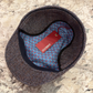 A herringbone pattern flat cap shown from above on a speckled stone surface, with a blue patterned interior lining and a red hangtag.