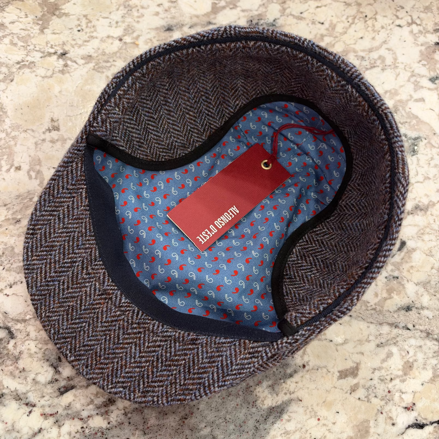 A herringbone pattern flat cap shown from above on a speckled stone surface, with a blue patterned interior lining and a red hangtag.