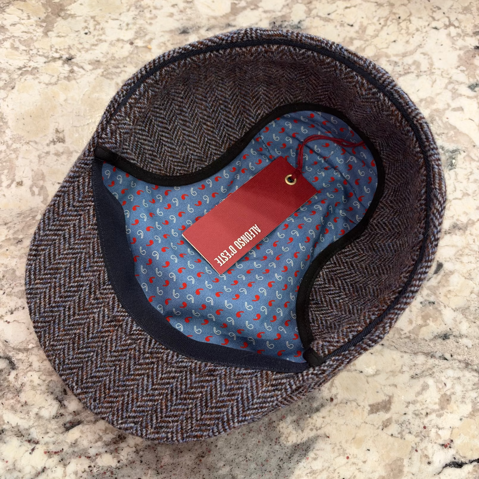 A herringbone pattern flat cap shown from above on a speckled stone surface, with a blue patterned interior lining and a red hangtag.