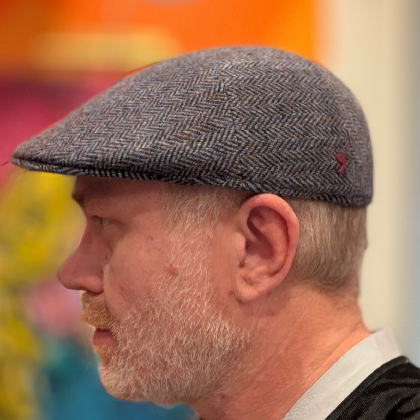 A man in profile wearing a gray herringbone flat cap, with a colorful blurred background.